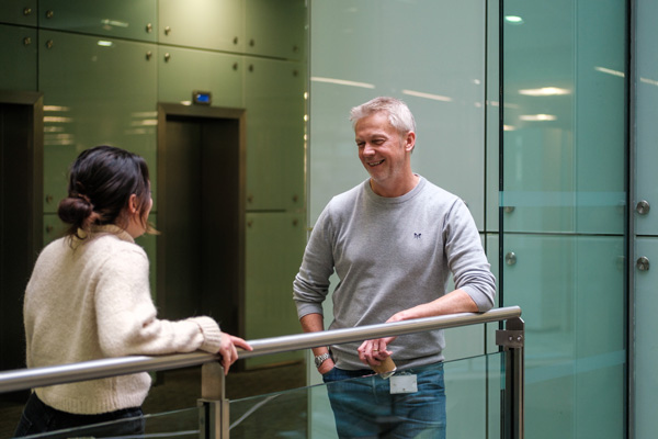 Two people standing and talking near a railing indoors, with glass walls and an elevator door in the background; one in a light sweater, the other in a grey top and blue jeans.