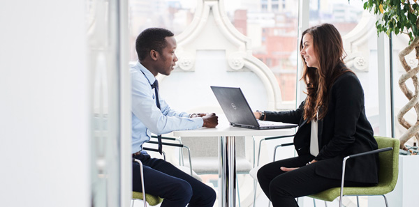 Two people in business attire sit across from each other at a round table; one uses a laptop.