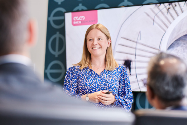 Person standing and speaking in front of an audience, with a large screen behind displaying a presentation featuring the 