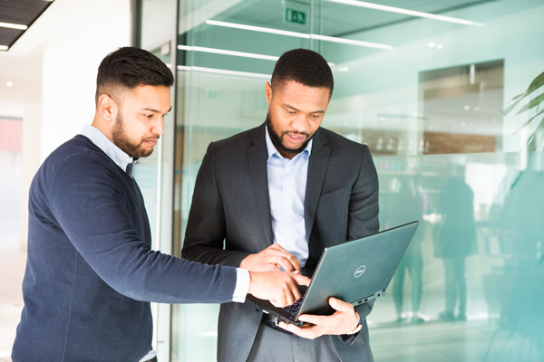 Two individuals in a modern office; one in a dark suit holding an open laptop, the other in a blue sweater pointing at the screen, with glass walls and reflections in the background.