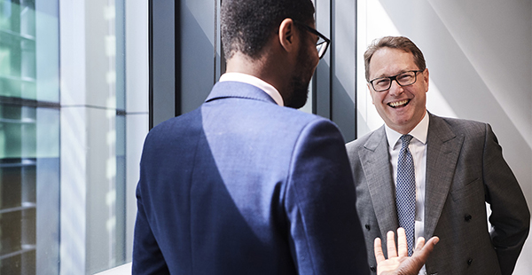 Two individuals in business attire conversing near large windows; one in a blue suit seen from behind, the other in a grey suit and tie facing forward.
