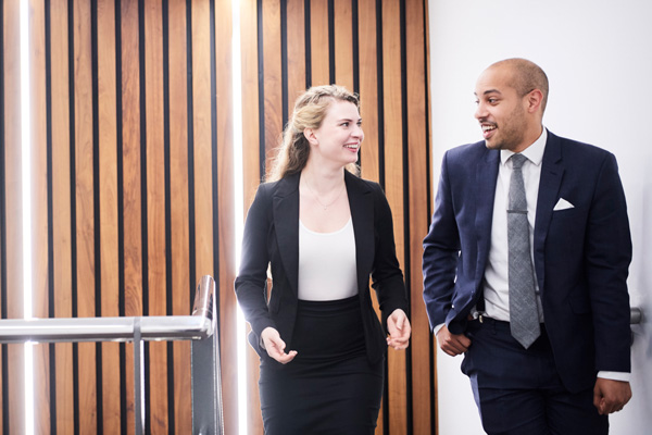 Two individuals in business attire walking and talking in a modern office with wooden panels and metal railing.