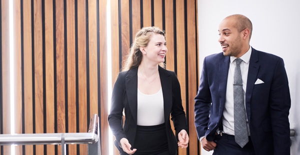 Two people in business attire walk past a wall with vertical wooden slats and a metal railing.