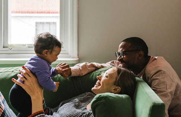 Two parents supporting their child standing on a green couch, with a window in the background.