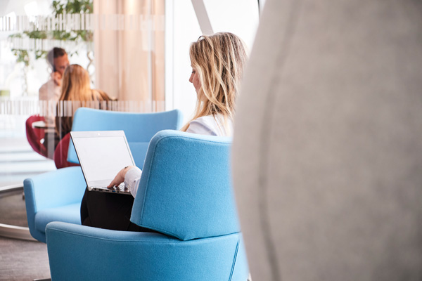 Person seated on a blue chair working on a laptop, with others in the background conversing in a modern lounge area with large windows and natural light.