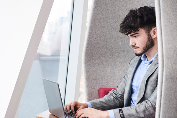 Person in a light blue shirt and grey blazer sits at a desk with a laptop in a modern workspace with large windows.
