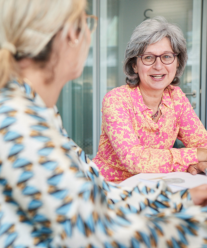 Two people seated at a table in conversation; one seen from behind in a blue and white patterned shirt, the other with grey hair in a pink and yellow patterned shirt.