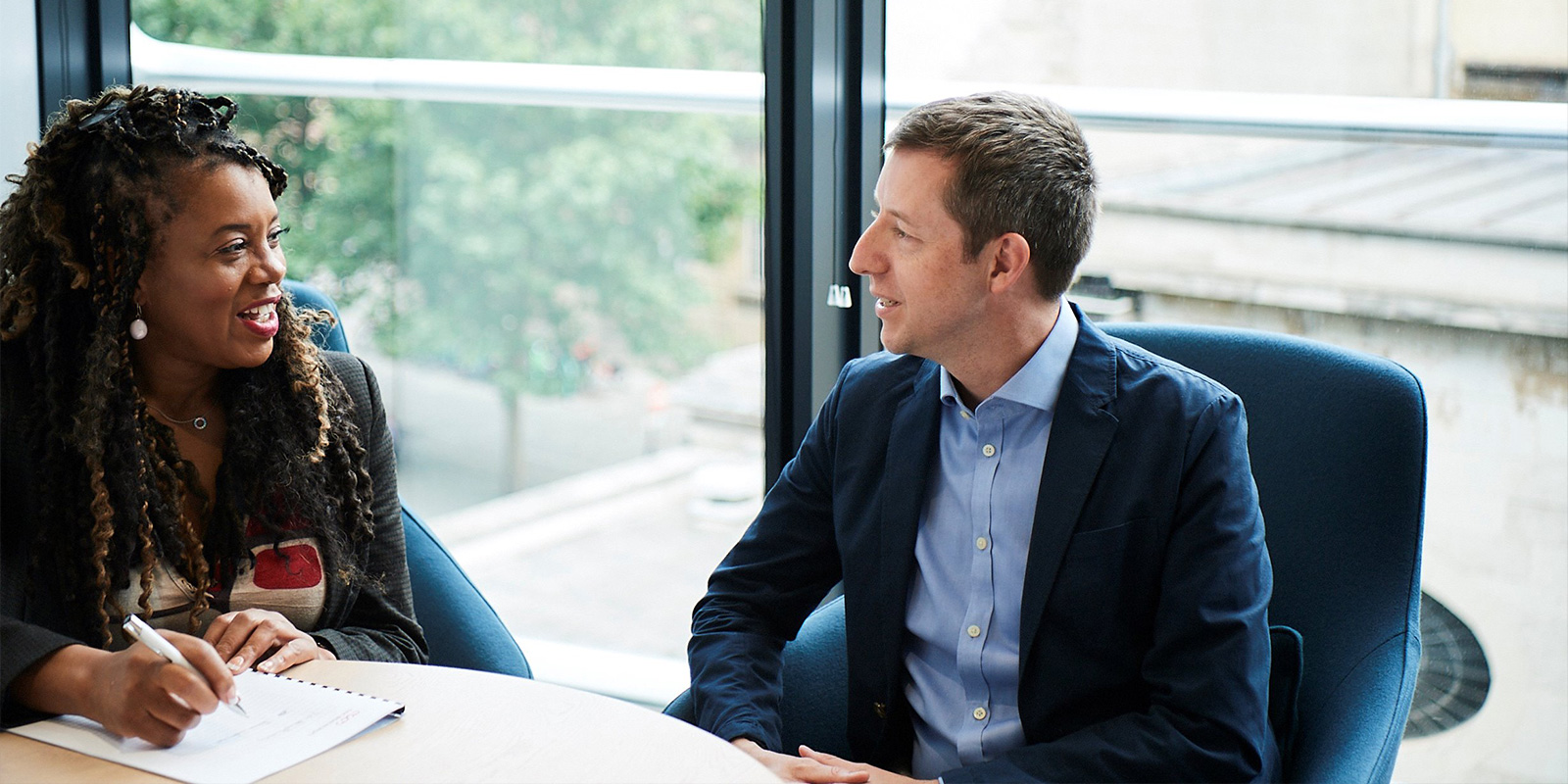 Two people at a round table in an office; one writing on a notepad, the other looking on, with blurred faces and large windows showing trees and buildings.