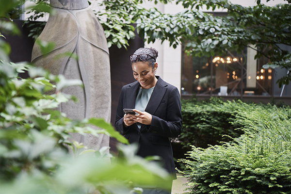 Person in formal attire using a smartphone in a garden, with a stone sculpture and building in the background.