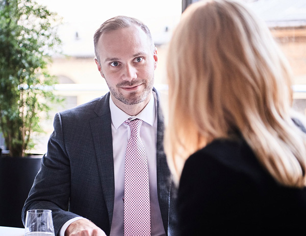 Two people seated across from each other at a table in a professional setting; one in a suit with light pink tie, the other with long blonde hair in dark clothing, with a glass of water, plants and windows in the background.
