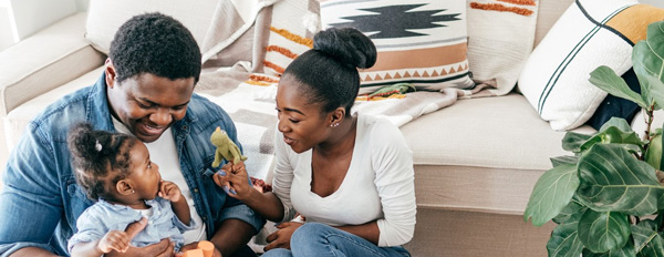 Man and woman sitting on the floor in a living room, engaging with a child using toys, with a couch and plant in the background.