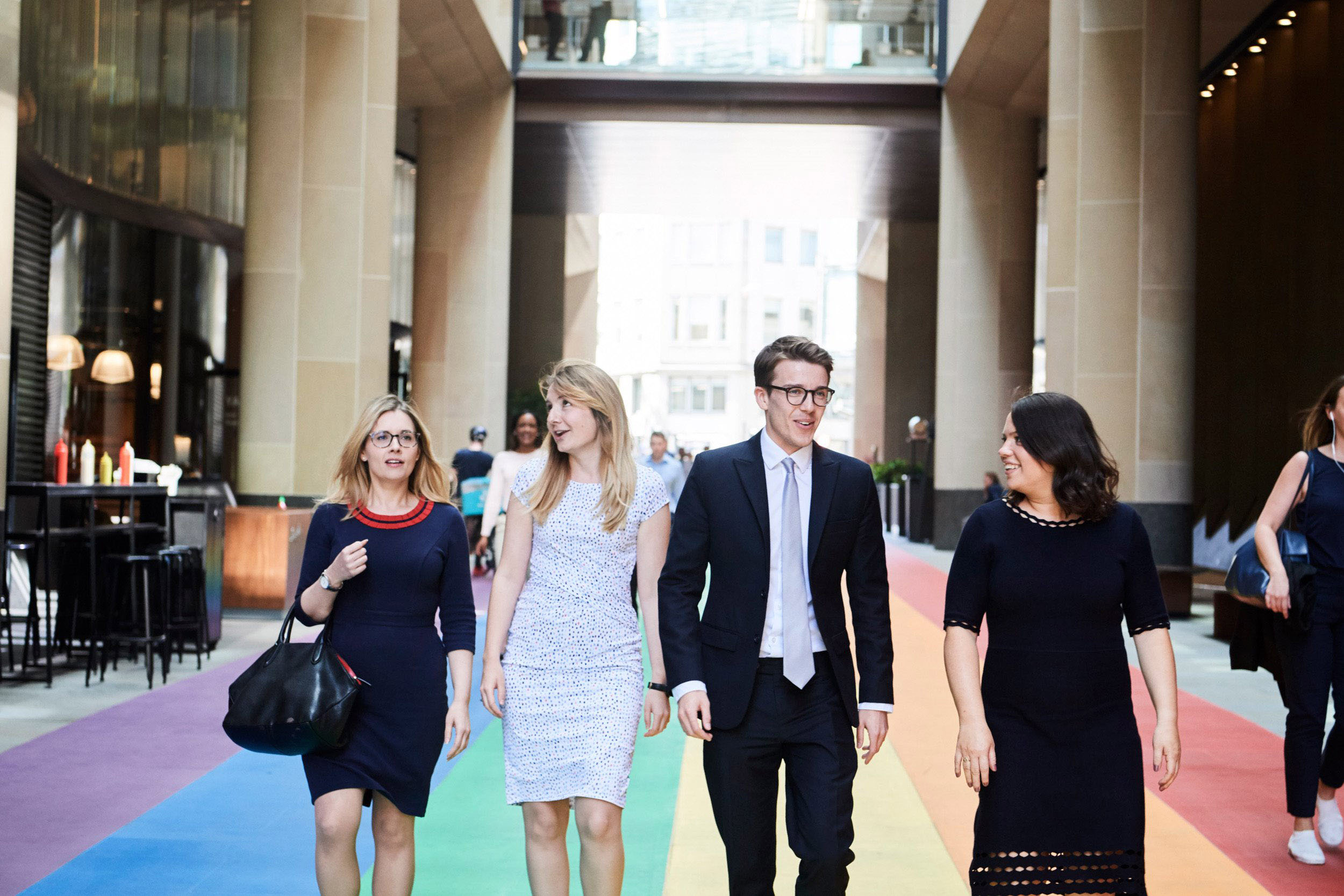 Four people walking on a rainbow-coloured pathway in an urban area, with tall buildings and a covered walkway in the background.