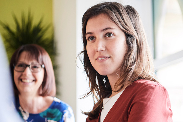 Two people are pictured indoors; one in a red top in the foreground, the other in a blue and white patterned top with a green plant and window in the background.