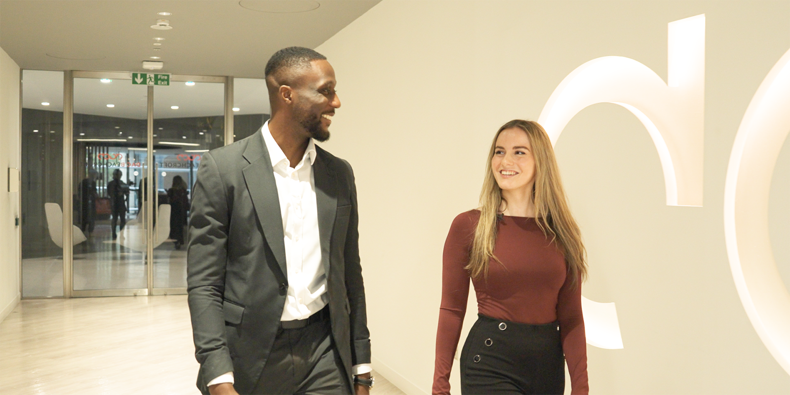 Two people are pictured indoors; one in a red top in the foreground, the other in a blue and white patterned top with a green plant and window in the background.